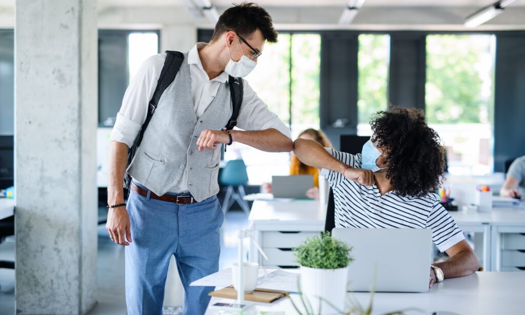Young people with face masks back at work in office after lockdown, greeting.